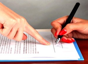 Closeup of paperwork and 2 hands, one pointing to sign, the other signing